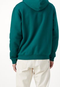 Grön hoodie visad bakifrån, med avslappnad passform, ribbade ärmslut och nederkant. Matchad med ljusbeiga byxor.