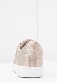 Beige metallisk sneaker med vit gummisula, med stickade detaljer och Reebok-logga på hälen. Slät textur, lågt snitt.