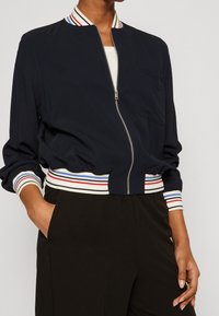 Námornická krátká bomber bunda se stojáčkem. Má bílé, červené a modré pruhované pružné manžety a lem. Přední zip a kapsy detail.