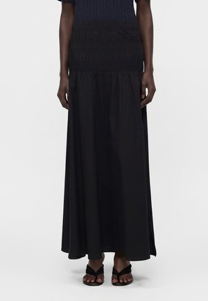 Γυναίκα που φοράει μακριά μαύρη φούστα με ψηλή μέση και ραφές στη μέση, καθώς και μαύρα σανδάλια ανοιχτού δαχτύλου.