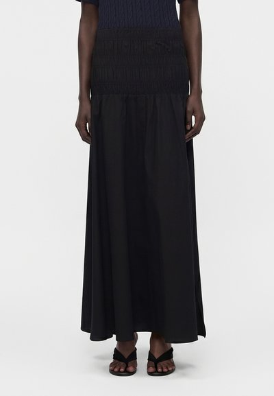 Femme portant une longue jupe noire taille haute avec des détails smockés à la taille et des sandales noires à bout ouvert.