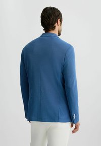 Blauer Blazer, Strukturstoff, taillierte Schnittführung, einzelner Schlitz, mit drei Knöpfen versehenen Manschetten, kombiniert mit hellfarbigen Hosen.