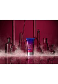 Crema retinolo avanzata in un tubo viola con cappuccio argentato, circondata da bottiglie di vetro da laboratorio, su uno sfondo rosa scuro.