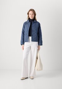 Quilted navy jacket with a structured collar and button closure, paired with fitted white pants and a cream tote bag. Smooth textures throughout.