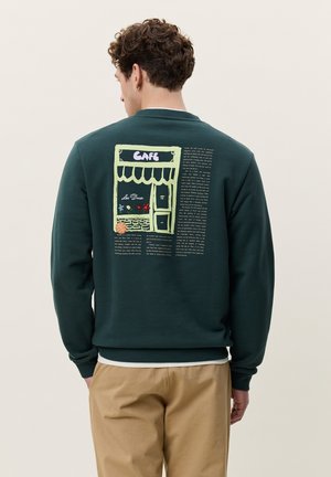 Homme portant un sweat-shirt vert foncé avec une illustration colorée de café et un texte au dos, associé à un pantalon beige, vu de dos.