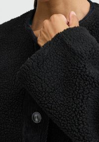 Svart fleecejacka med texturerad tyg, rund hals och breda ärmar. Har en svart knappstängning och kontrasterande ribbade detaljer.