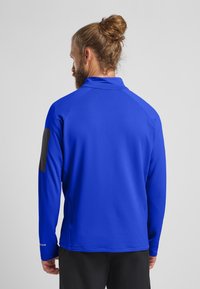 Blaues langärmliges Sportoberteil aus strukturiertem Stoff, mit schwarzer Akzentfarbe auf der linken Schulter und einem Stehkragen, Ansicht von hinten.