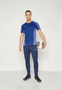 Sinine spordusärk lühikeste varrukatega, lilla aktsentidega ja Nike logo, mille juurde on kombineeritud tumedast sinisest sobivustest püksid; mudel hoiab jalgpalli.