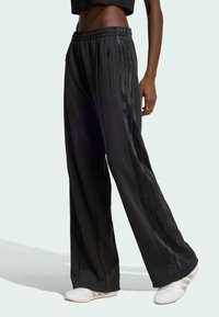 Black wide-leg pants with an elastic waistband, featuring glossy side stripes and zip pockets, paired with white athletic shoes.