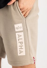 Beige sporta šorti, izgatavoti no mīksta auduma, ar sānu kabatu un izteiksmīgu baltu "ALPHA" logo ar akcentu detaļu.