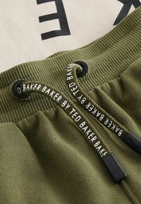 Olivové teplákové nohavice s rebrovaným pásom, disponujúce čiernym šnúrkou s bielym textom "Baker by Ted Baker." Hladká textúra látky.