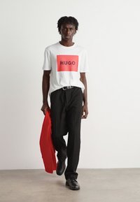 Hvit t-skjorte med en rød rektangulær logo som sier "HUGO," kombinert med svarte bukser. Modellen holder en rød blazer og har på seg svarte sko.