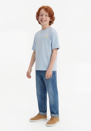 Smilende rødhåret gutt iført lyseblå t-skjorte, opprullede blå jeans og beige joggesko, står mot en ensfarget hvit bakgrunn.