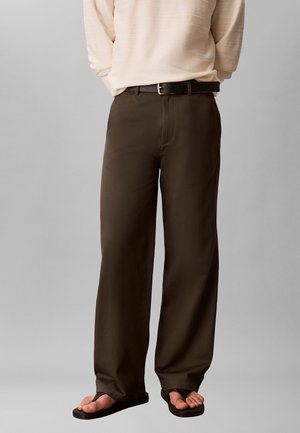 Osoba nosząca ciemnobrązowe spodnie z szerokimi nogawkami, czarny pasek, beżową bluzkę z długim rękawem i czarne klapki na jednolitym tle.