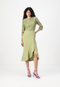 Robe vert olive avec un corsage en dentelle, des manches trois-quarts et un ourlet volé, associée à des talons mitaines violets.