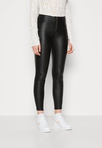 Juodos dirbtinės odos legging kelnės su aukštu liemeniu ir priekinėmis užtrauktuko detalėmis, prigludusio silueto, derinamos su baltais sportbačiais su rožine swoosh detale.