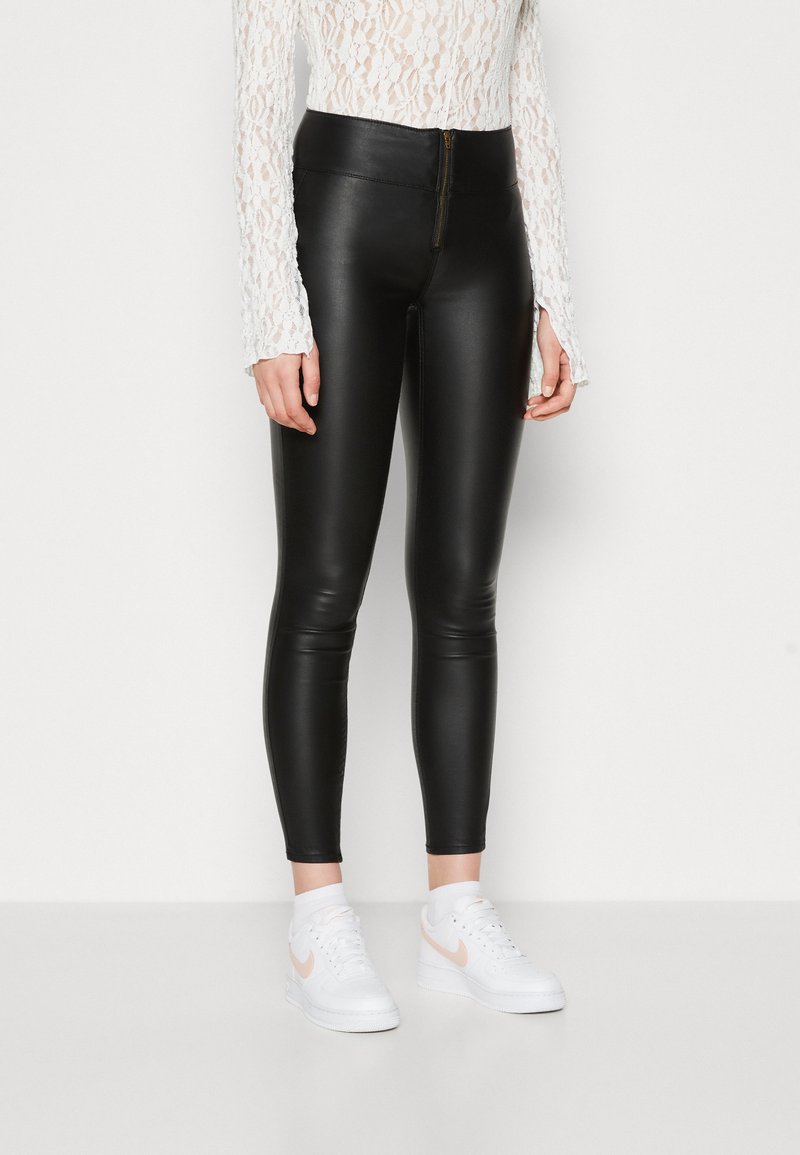 Juodos dirbtinės odos legging kelnės su aukštu liemeniu ir priekinėmis užtrauktuko detalėmis, prigludusio silueto, derinamos su baltais sportbačiais su rožine swoosh detale.