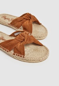 Des sandales en daim brun à enfiler présentent un design noué et une semelle en jute tissée. Les accents incluent un bord cousu et une semelle intérieure texturée.