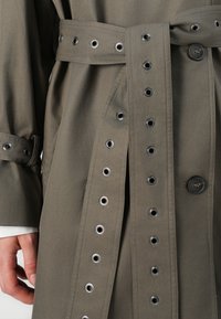 Olive rohelise värvi trench coat, millel on suured silmad nööbisuletuse ja vöö ümber; omab klassikalist kuju ja siledat kangast tekstuuri.