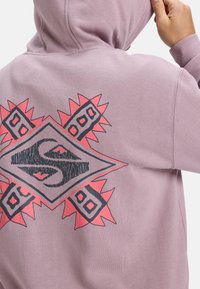 Sudadera morada con un diseño gráfico en la parte posterior que presenta una forma de diamante y acentos rojos llamativos, incluyendo patrones y formas abstractas.