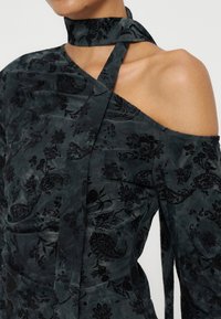 Haut en velours vert foncé avec motifs floraux noirs, design asymétrique à une épaule et un lien assorti noué autour du cou.