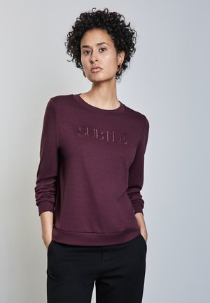 Felpa bordeaux con maniche lunghe, collo rotondo e testo impresso "SUBTLE" sul davanti. Tessuto morbido, vestibilità casual, abbinata a pantaloni neri.