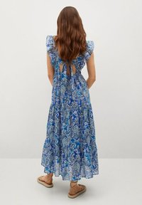 Blaues Paisley-Maxikleid mit Rüschenschößen und ausgeschnittenem Rückendetail. Weicher Stoff, fließendes Design, knöchellang, kombiniert mit flachen Sandalen.