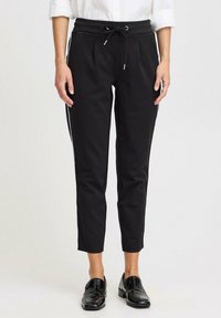 Mustat housut, joissa on kiristysnauha vyötäröllä, kapeaksi kapeneva malli ja sivuviirut. Varustettu kahdella etutaskulla ja sileällä pinnalla. Yhdistetty kiiltäviin mustiin loafer-kenkiin.