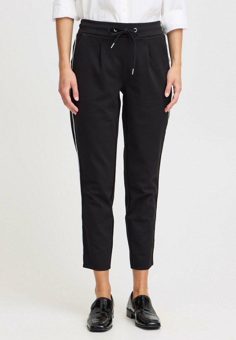 Mustat housut, joissa on kiristysnauha vyötäröllä, kapeaksi kapeneva malli ja sivuviirut. Varustettu kahdella etutaskulla ja sileällä pinnalla. Yhdistetty kiiltäviin mustiin loafer-kenkiin.