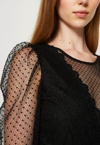 Abito nero con motivo in pizzo floreale, maniche a sbuffo trasparenti con pois neri, dettaglio del colletto a scallop, tessuto testurizzato.