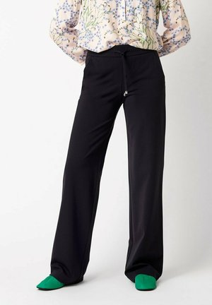 Woman wearing loose black pants with drawstring and green pointed shoes, paired with a floral long-sleeve blouse.