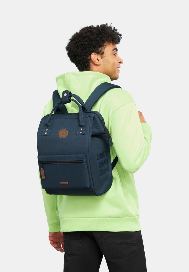 Zaino in tessuto blu con manici superiori e spallacci regolabili, dotato di una tasca frontale con zip e dettaglio logo. Esterno liscio e forma strutturata.