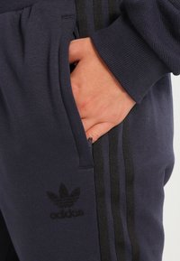 Námořnické tepláky s černými pruhy, vyrobené z měkké tkaniny. Obsahují boční kapsu a černé vyšívané logo na stehně.