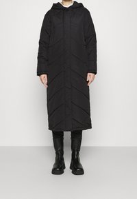 Black puffer coat with a hood, featuring a chevron quilted pattern, long length, and front button closure. Worn with black knee-high boots.