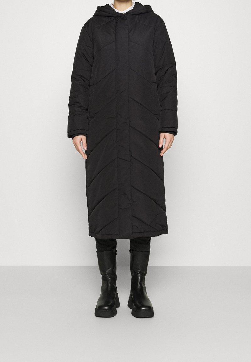 Black puffer coat with a hood, featuring a chevron quilted pattern, long length, and front button closure. Worn with black knee-high boots.