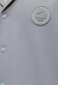 Chemise gris clair boutonnée avec un patch brodé rond indiquant « BOSS GRAND SLAM » et un motif de balle de tennis sur la poitrine.