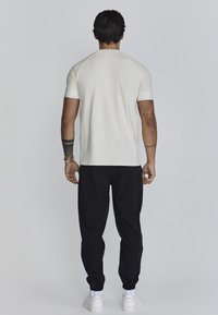 Vaaleanbeige lyhythihainen t-paita pyöreällä pääntiellä, yhdistetty mustiin jogginghousuihin. Malli kääntynyt pois päin, minimalistiinen muotoilu, ilman näkyviä kuvioita.