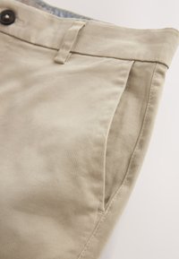 Pantalon en coton beige avec poche latérale avant, passants de ceinture et bouton noir visible à la taille.