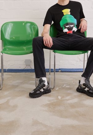 Personne portant un pantalon noir, un t-shirt cartoon noir et vert, et des baskets noires assise, les jambes écartées, sur une chaise verte à côté d'une chaise identique vide.