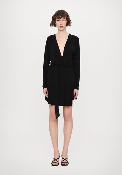 Svart omlottklänning med långa ärmar, djup V-ringning och skärp i midjan. Mjuk tyg, utsvängd fåll, bärs med svarta remsandaler.