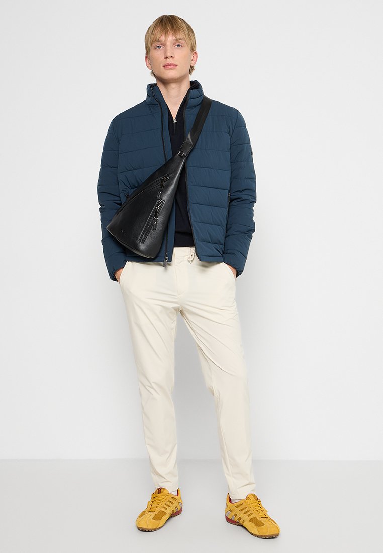 Jonge man in een blauwe gewatteerde jas, crèmekleurige broek, gele sneakers en een zwarte schoudertas, staande tegen een lichte achtergrond.