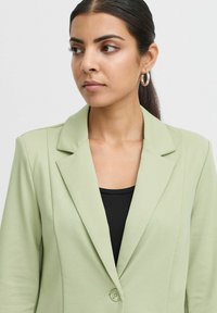 Světle zelený blazer se strukturovaným designem, klopami a jedním knoflíkem. Ušitý z hladké látky. Nosený přes černý top.
