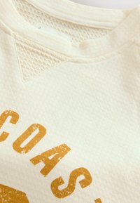 Beige texturerad sweatshirt med rund halsringning, utrustad med ett upphöjt mönster på axlarna och en tryckt guldfärgad grafisk design.