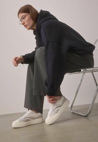 Sneaker grigie chiare e bianche con suola spessa, caratterizzate da un accento nero curvato, abbinate a pantaloni grigi e una felpa blu navy.