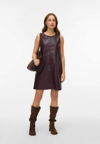 Vestido sin mangas de cuero sintético color burdeos con corte recto, dobladillo a la altura de la rodilla y detalles mínimos de costura. La modelo lleva un bolso marrón.