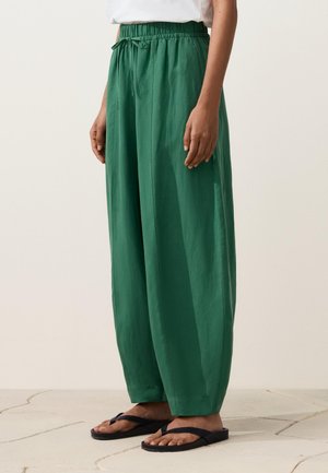 Osoba odjevena u široke zelene hlače s vezicama, bijelu majicu i crne japanke stoji na svijetlom pločastom podu.