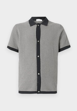 Πολυεστερικό πόλο μπλουζάκι κοντό μανίκι σε μαύρο και άσπρο ριγέ ύφασμα, με μαύρο γιακά και μπροστινό κλείσιμο με κουμπιά διαθέσιμα σε ασημένια απόχρωση.