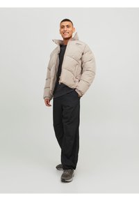 Chaqueta acolchada beige con un alto cuello, diseño acolchado, cremallera frontal y logo de marca. Lleva puesta sobre una camisa negra y combinada con pantalones negros.