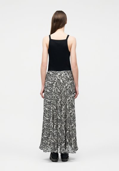 Frau mit langen braunen Haaren, die ein schwarzes Tanktop, einen langen Rock mit schwarz-weißem Zebramuster und schwarze Turnschuhe trägt, steht mit dem Rücken zum Betrachter vor einem weißen Hintergrund.