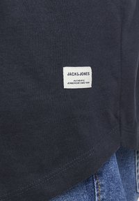 Camiseta de algodón azul oscuro con una etiqueta blanca que dice "JACK & JONES" y "AUTHENTIC JEANSWEAR SINCE 1990" cerca del dobladillo.